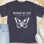 Dark grey t-shirt with white text "BECAUSE HE LIVES i can face tomorrow" and a butterfly design.